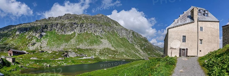 Panoramic photo view to the right left side of Ospizio Hospiz Herberge San Gottardo St. Gotthard built in the 13th century from 1230 with small entrance door main entrance to chapel Cappella di San Gottardo on pass summit of Gotthard Pass, left mountain lake Lago di Monti, Swiss Alps, Airolo, Canton Ticino, Ticino, Switzerland