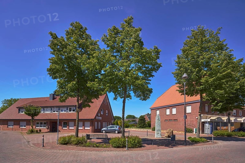 Street, bollards, trees, bench, 1000 year fountain, lantern, clinker brick pavement, concrete paving stone street, shady, pavement, clinker brick building, hedges, blue sky, cloudless, intersection of Hauptstraße with Nonnenpfad and In den Gärten, Alfhausen, Osnabrück district, Lower Saxony, Germany