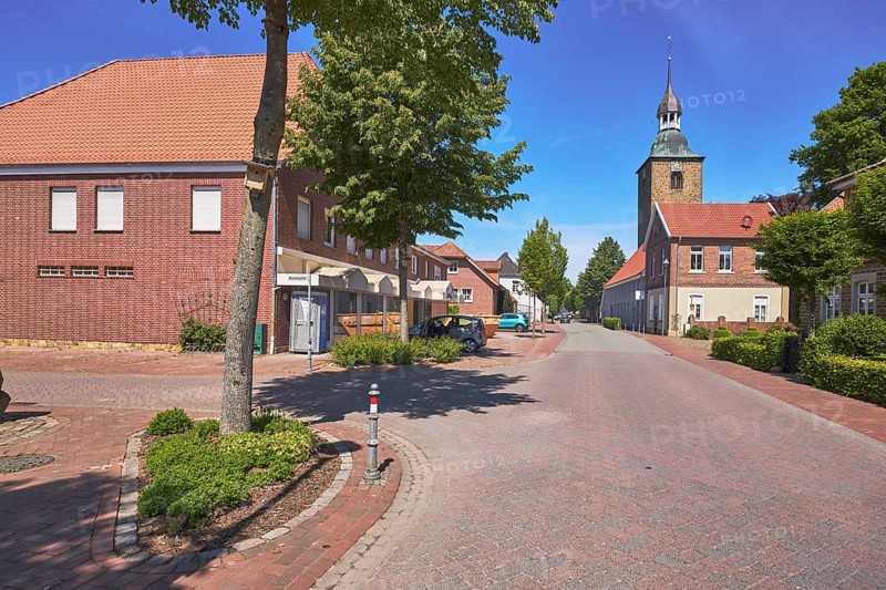 Street, general development, clinker brick building, pointed roof with red roof tiles, church tower of St. Johannis, trees, clinker brick pavement, asphalt road, pavement, blue sky, cirrostratus clouds, intersection of Hauptstraße and Nonnenpfad, Alfhausen, district of Osnabrück, Lower Saxony, Germany