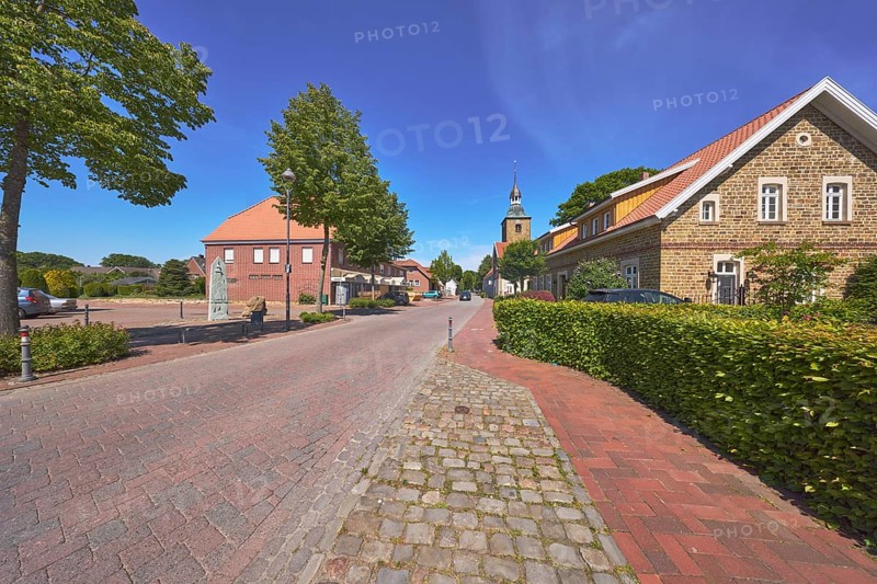 Historic brick building, pointed roof with red roof tiles, church tower of St. Johannis, hedge, trees, clinker pavement, street, pavement, blue sky, cirrostratus clouds, main street, Alfhausen, district of Osnabrück, Lower Saxony, Germany