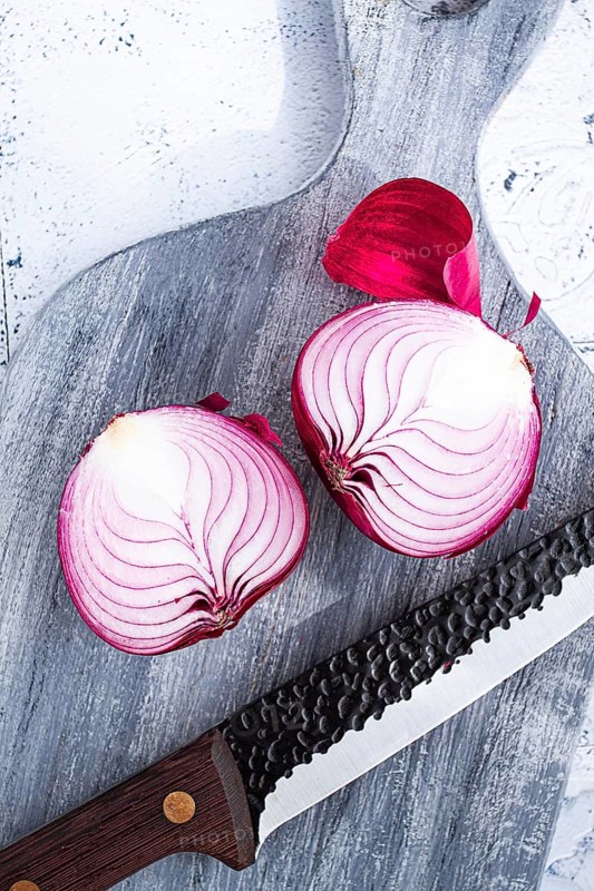 A vibrant red onion is cut in half, on a rustic wooden chopping board beside a sharp knife, ready for cooking