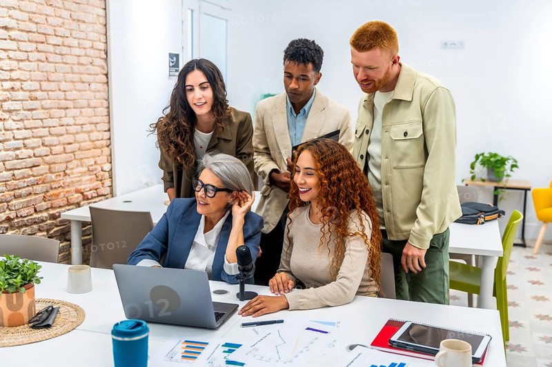 Diverse business team working together in a modern office, grouping around a laptop and microphone, discussing data charts and sharing ideas during a casual meeting