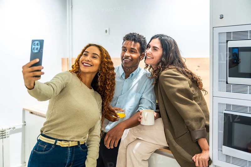 Group of diverse colleagues enjoying a coffee break in the office kitchen, smiling and posing together for a selfie using a smartphone, showing friendship and teamwork