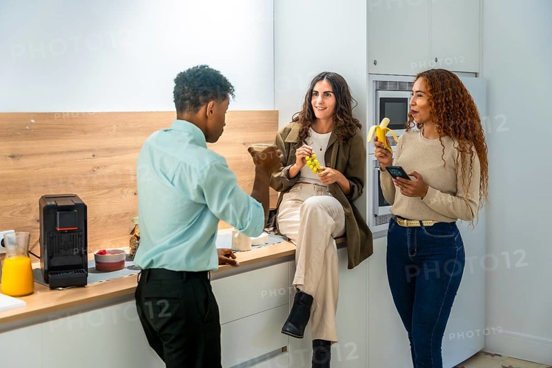 Diverse colleagues chatting and smiling during a casual office kitchen break, two women holding fruit and a smartphone while a male coworker talks, relaxed teamwork vibe