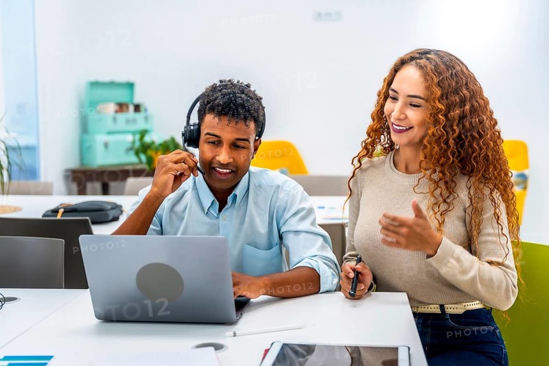 Diverse colleagues at an office desk collaborate as a man with a headset uses a laptop while a woman supports and communicates, smiling and providing customer service and technical help