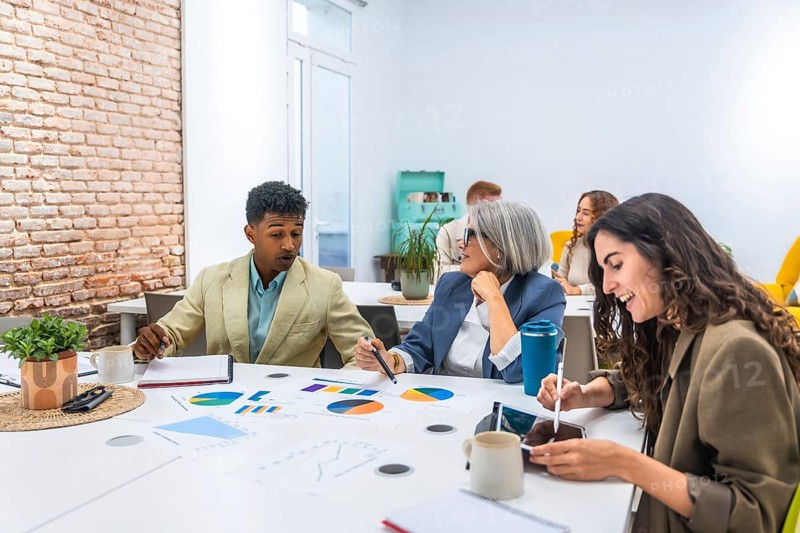 Multiracial business professionals discussing colorful charts and data analytics during a collaborative meeting, working together on a project in a bright, contemporary office environment