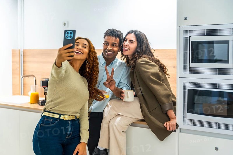 Diverse coworkers cheerfully taking a selfie on a smartphone during a relaxed office kitchen break, sharing laughs and casual bonding over coffee and snacks