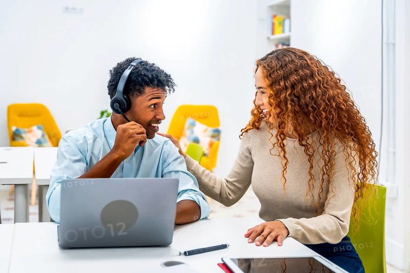 Two diverse colleagues in a modern office setup, a man wearing a headset using a laptop while a woman offers support and guidance, highlighting teamwork and effective communication