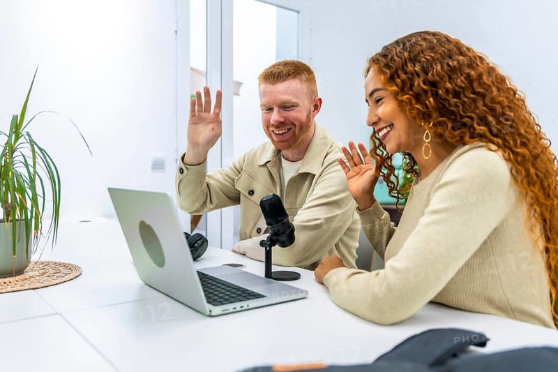 Diverse young adult man and woman smiling and waving while actively recording a podcast or participating in a video call event from home, using a laptop, microphone, and headphones