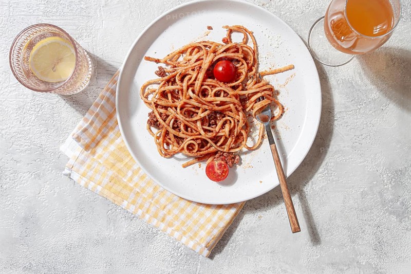 A plate of freshly cooked pasta sits on a textured surface. The pasta is topped with cherry tomatoes and herbs, accompanied by a glass of refreshing drink and a yellow checkered napkin