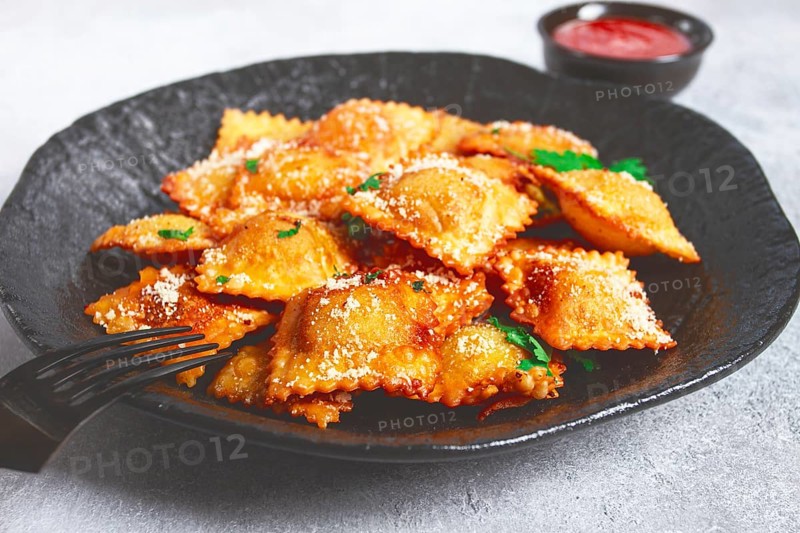 Fried ravioli with parmesan cheese, served with red sauce and garnished with herbs, on a black plate, no people