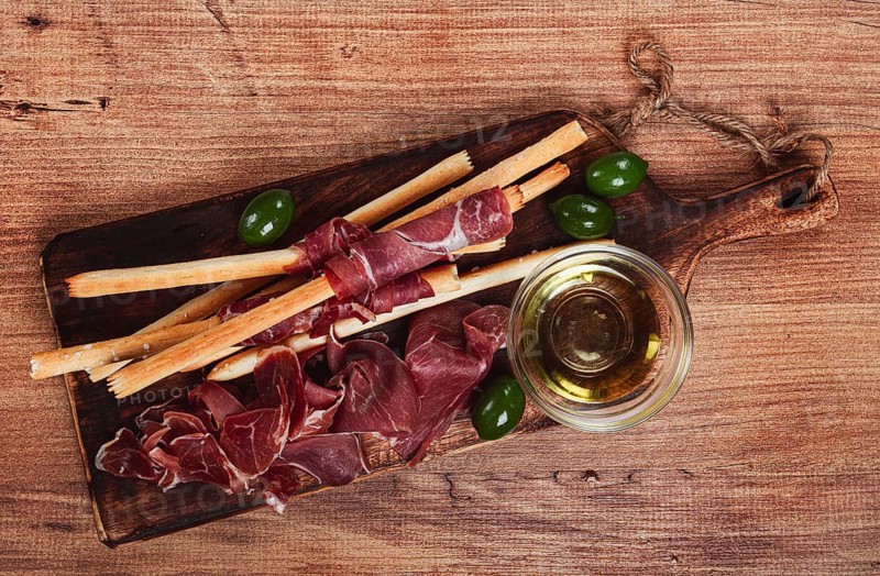 An appetizer of prosciutto, grissini and olives, served on a wooden board, with olive oil