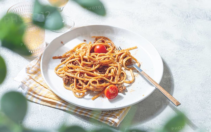 A plate of perfectly cooked spaghetti is topped with rich sauce and fresh tomatoes. Next to it, a glass of cold drink sits on a light tablecloth, creating a cozy dining scene
