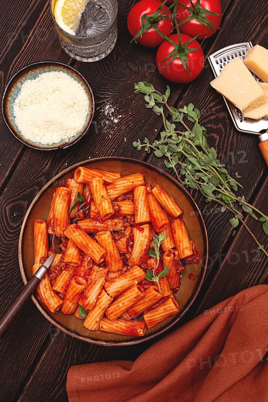 Rigatoni pasta with meat sauce and parmesan cheese on a dark wood table with ingredients, top view