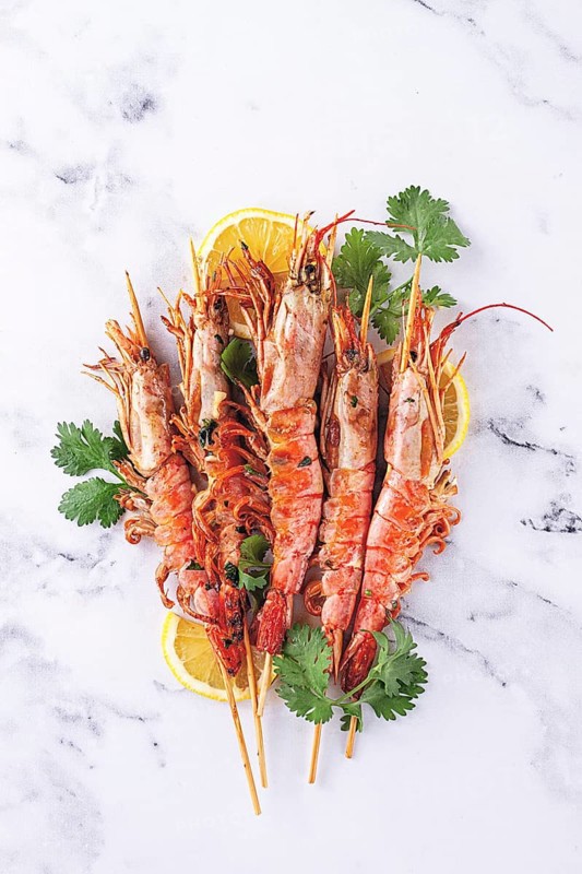 Shrimps on skewers, Grilled, spread on a marble surface, garnished with lemon slices and fresh herbs. view from above