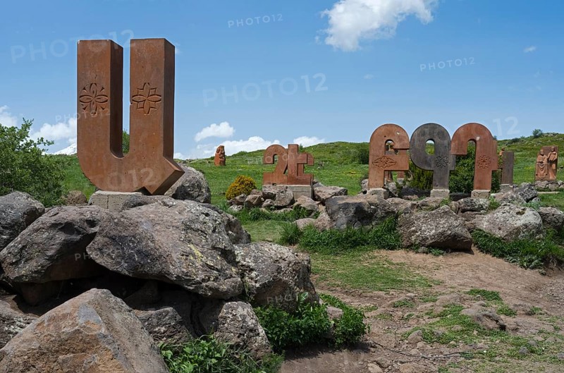 Row of stone columns with motifs, integrated into a rocky, green landscape under a blue sky, monument to the Armenian alphabet, Aparan, Aragazotn Province, Armenia