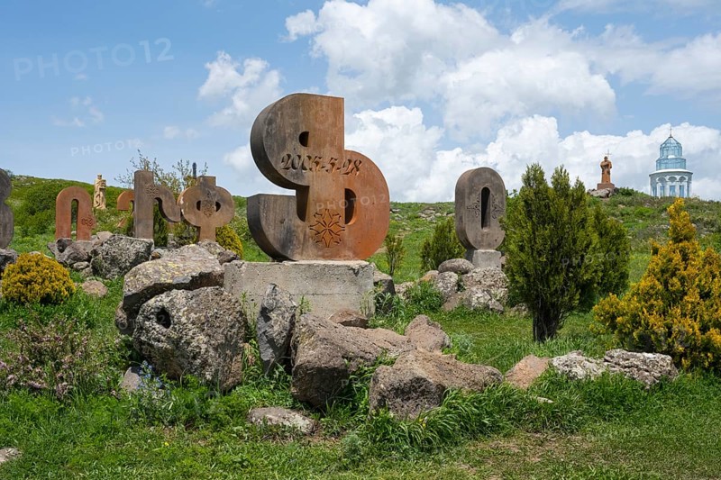 Stone sculptures in the shape of letters on a green meadow, surrounded by rocks and bushes under a cloudy sky, monument to the Armenian alphabet, Aparan, Aragazotn Province, Armenia