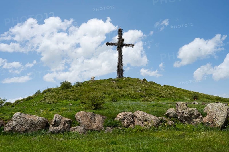 Metal cross on a green hill under a blue sky, surrounded by large stones, Monument to the Holy Cross, consisting of many small metal crosses, Aparan, Aragazotn, Armenia