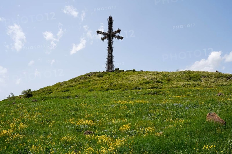 Metal cross on a green hill under a cloudy blue sky, surrounded by grass and yellow flowers, Monument of the Holy Cross, consisting of many small metal crosses, Aparan, Aragazotn Province, Armenia
