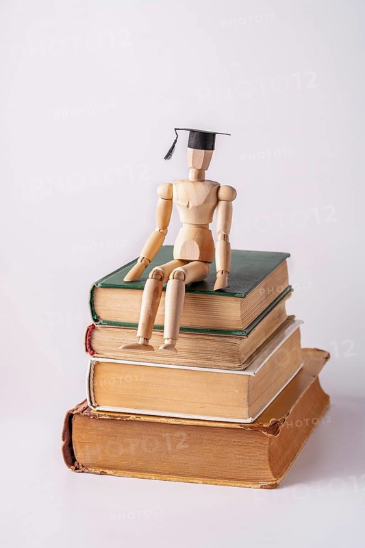 A wooden man in a student cap sits on a stack of textbooks on a white background