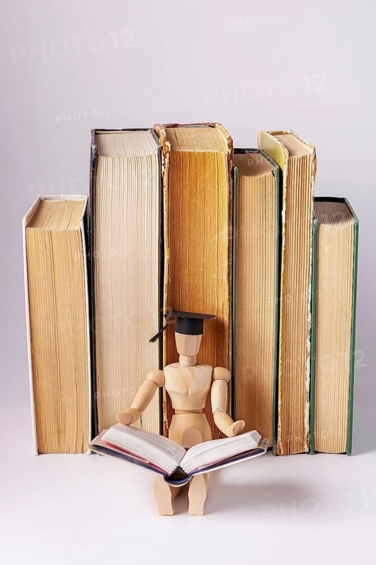A wooden man in a student cap sits against a background of a stack of textbooks and reads a book, space for text