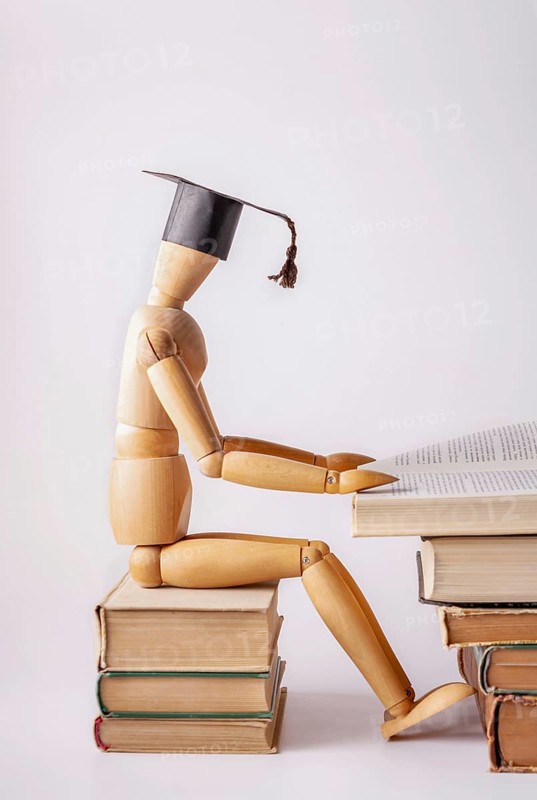 A wooden man in a student cap sits on a stack of textbooks and reads a book