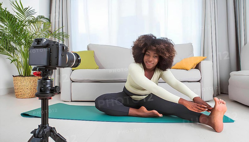A woman is doing a yoga pose in front of a camera. The camera is on a tripod and is recording her pose. The woman is smiling and she is enjoying herself. The room is decorated with a couch, AI generated