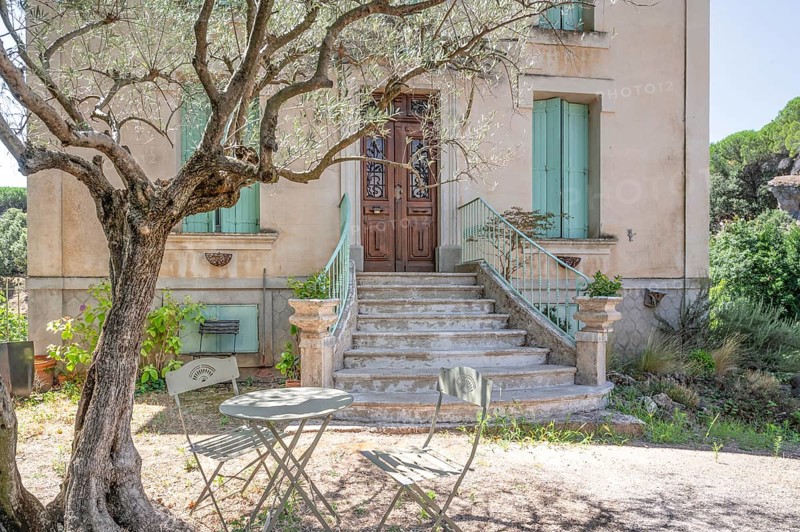 Typical Midi architecture in the south of France: a rustic stone staircase with ornate banisters leads to a dark brown wooden door of an ochre-coloured house, while a small bistro table waits in the shade of an olive tree: Moureze, Herault, Occitania, France