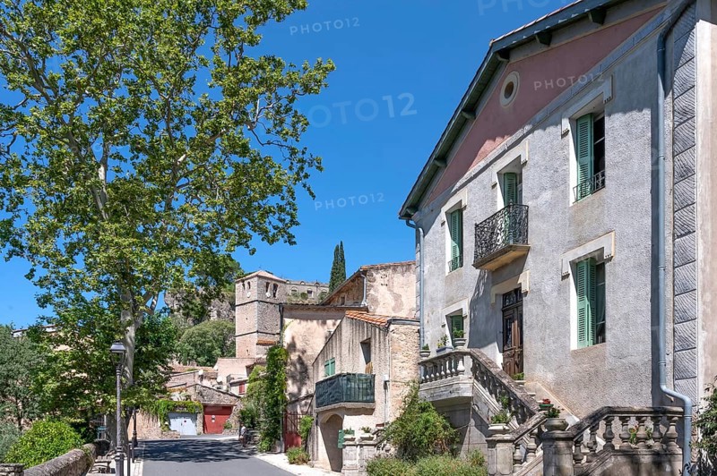 The sunlit village street of Moureze is lined with traditional stone houses with reddish-brown garage doors, while the medieval bell tower and the craggy cliffs of the Cirque de Moureze loom in the background: Moureze, Grand Site Salagou-Cirque de Moureze, Herault, Occitania, France