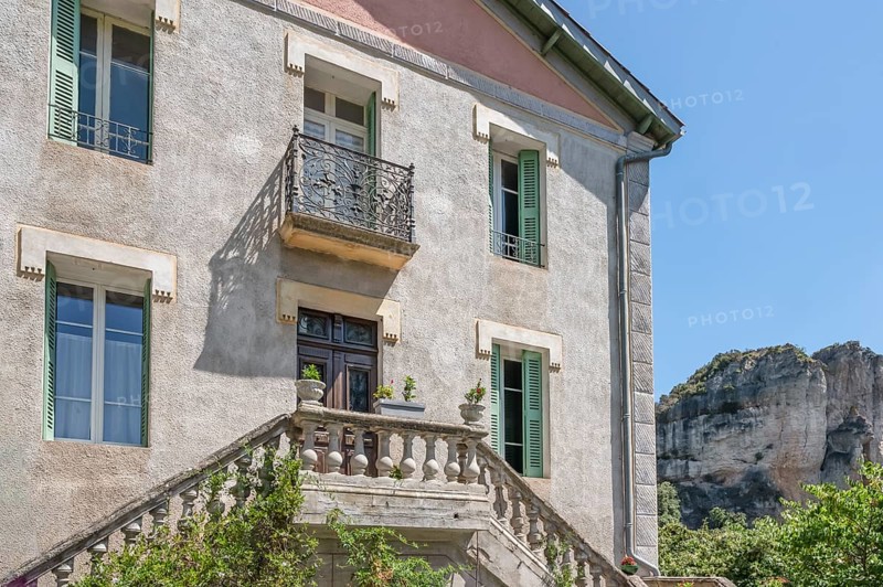 Typical Midi village architecture with stone balustrade and wrought-iron balcony of a light grey building with green shutters, while the bizarre, eroded dolomite cliffs of the Cirque de Moureze tower in the background: Moureze, Herault, Occitania, France