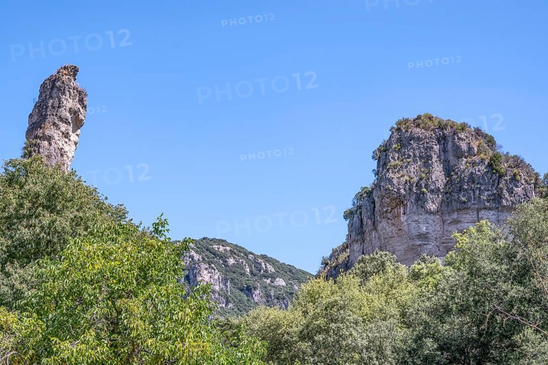 Two prominent dolomite cliffs formed by erosion - a towering peak on the left and a massive rock face on the right - above lush green vegetation under a blue sky, typical of the Cirque de Moureze: Moureze, Herault, Occitania, France