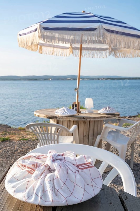 A plate wrapped in a red and white chequered tea towel lies on a rustic wooden table in the foreground at the beach restaurant Guinguette a la Bonne Franquette, with a blurred view of the glistening waters of the Etang de Thau, a round dining table with parasol and the busy coastline in the background, Sete, Herault, Occitania, France