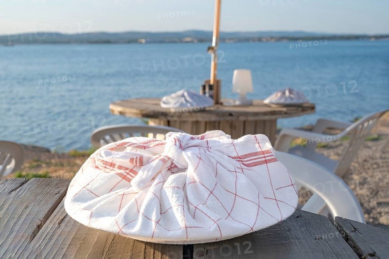 A plate wrapped in a red and white chequered tea towel lies on a rustic wooden table in the foreground of the beach restaurant Guinguette a la Bonne Franquette, with a blurred view of the glistening waters of the Etang de Thau and the lively coastline in the background, Sete, Herault, Occitania, France