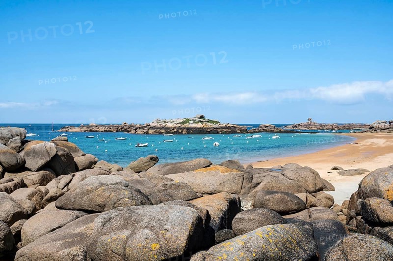 The wide bay of Tregastel on the Cote de Granit Rose shows the turquoise blue sea with white sailing boats and the characteristic round pink granite rocks, framed by large boulders in the foreground, Tregastel, Cotes-d'Armor department, Brittany region, France