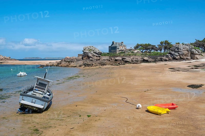 On the harbour beach of Tregastel on the Cote de Granit Rose, small boats lie on the light sand at low tide against the backdrop of the iconic pink granite rocks and Breton houses, Tregastel, Cotes-d'Armor department, Brittany region, France