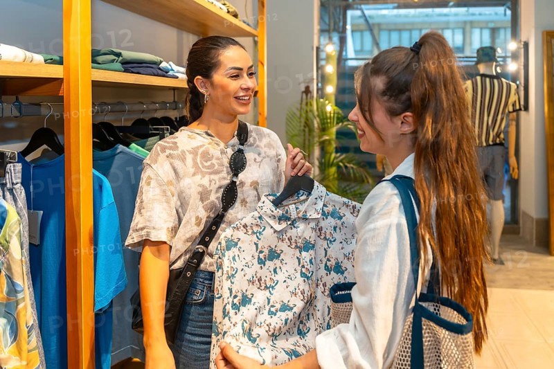 Two women interacting, choosing clothes in a store, one showing a shirt to the other, smiling and happy