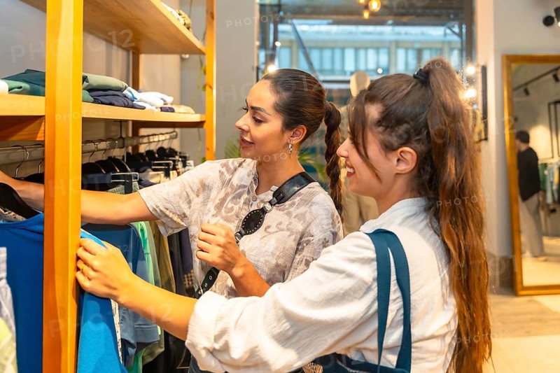 Two young women looking and choosing clothes in a fashion store, the shop assistant is helping a customer