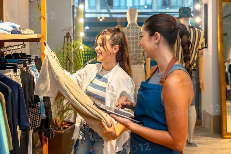 Smiling shop assistant holding tablet and showing trousers information to a happy customer in a clothing store