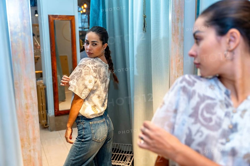 Young woman trying on new clothes looking at herself in a mirror in a dressing room of a fashion store