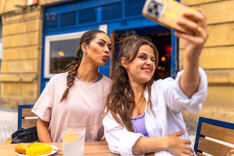 Two young women are taking a selfie with a smartphone while sitting at an outdoor table, enjoying breakfast