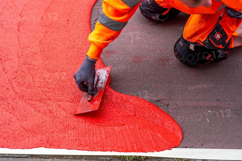 Application of red road surfacing on a cycle lane, inner-city road, so-called cold plastic surfacing, in the colour traffic red, RAL3020, the coloured marking is intended to attract more attention from motorists for cyclists, on cycle paths along roads and at junctions, roundabouts, Essen, North Rhine-Westphalia, Germany