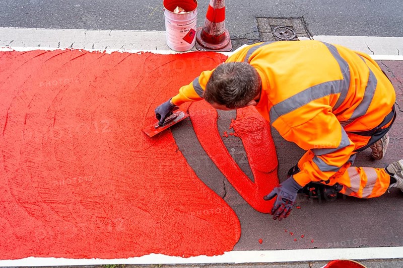 Application of red road surfacing on a cycle lane, inner-city road, so-called cold plastic surfacing, in the colour traffic red, RAL3020, the coloured marking is intended to attract more attention from motorists for cyclists, on cycle paths along roads and at junctions, roundabouts, Essen, North Rhine-Westphalia, Germany