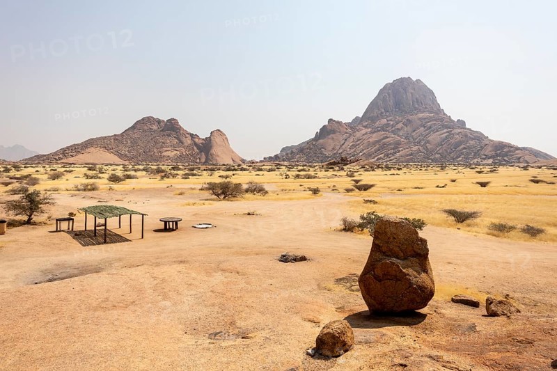 Granite peaks and rugged desert landscape at Spitzkoppe Community Campsite, Namibia. Golden grassland with scattered acacia trees contrasts against dramatic rocky mountains under a clear sky