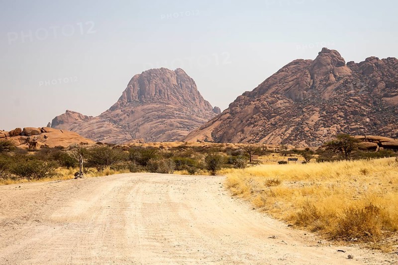 Granite peaks and rugged desert landscape at Spitzkoppe Community Campsite, Namibia. Golden grassland with scattered acacia trees contrasts against dramatic rocky mountains under a clear sky