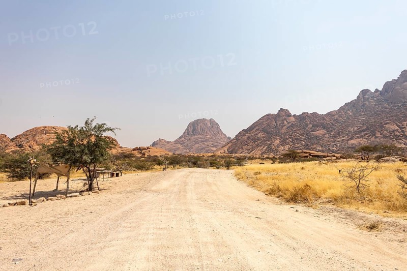 Granite peaks and rugged desert landscape at Spitzkoppe Community Campsite, Namibia. Golden grassland with scattered acacia trees contrasts against dramatic rocky mountains under a clear sky
