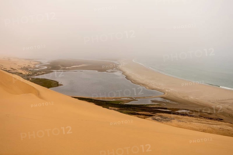 Golden dunes and the Atlantic Ocean near Namib-Naukluft Park, Namibia. A lagoon and shoreline fade into misty, cloudy weather, creating a dramatic desert-sea landscape
