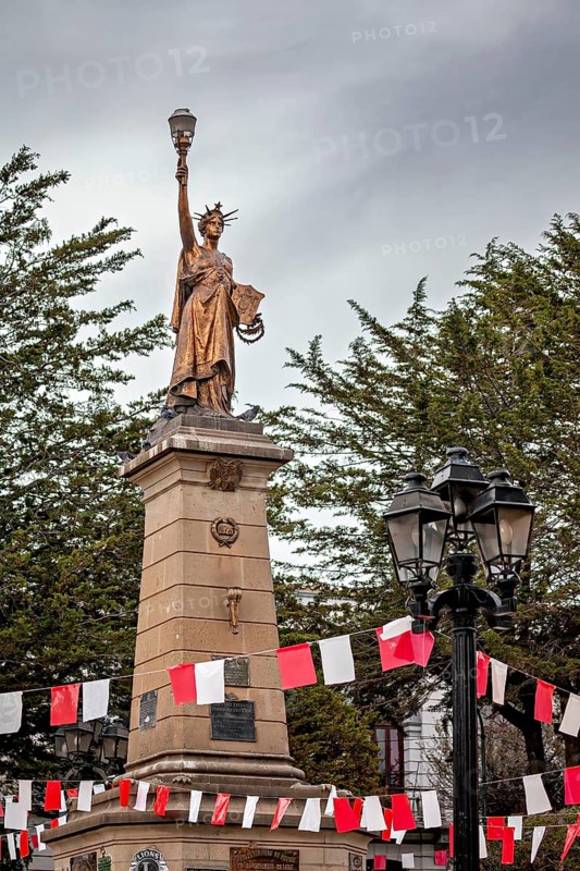 Historical statue with lantern on a high pedestal, surrounded by red and white flags and trees under a cloudy sky, a monument similar to the Statue of Liberty in the city of Potosi in Bolivia