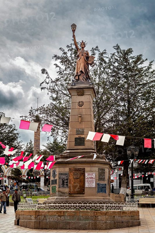 Statue on a stone pedestal in a plaza with red and white flags and trees, under a cloudy sky, Monument of a statue that looks like the Statue of Liberty in Potosi in Bolivia