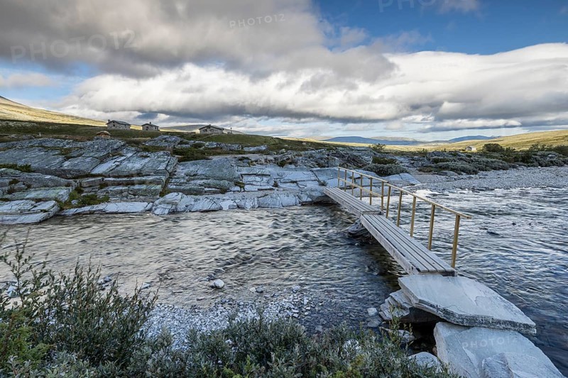 A small wooden bridge crosses a wide river, surrounded by rocks and sparse vegetation in the background, Peer Gynt hytta, Rondane National Park, Otta, Innlandet, Norway