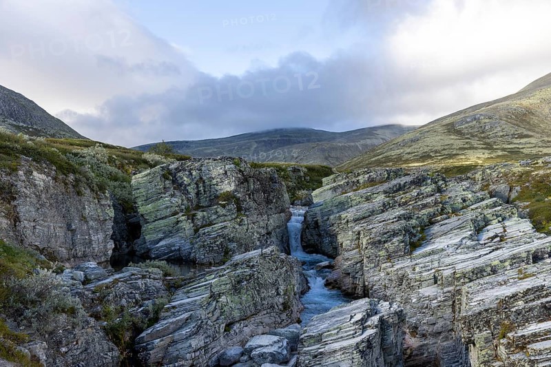 Rocks and a gorge through which a stream flows with a waterfall in the background, surrounded by mountains and clouds, Peer Gynt hytta, Rondane National Park, Otta, Innlandet, Norway